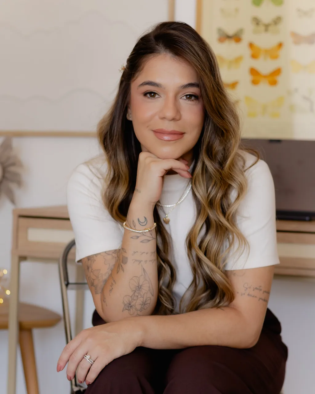 Andressa Petrolini Liani com o queixo apoiado na mão e expressão serena, em seu espaço de trabalho com um quadro de borboletas ao fundo