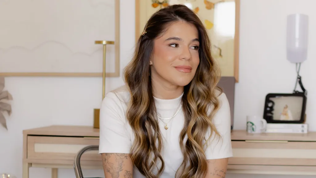 Andressa Petrolini Liani, da Agência Pettra, sentada em uma cadeira em um ambiente claro, com mesa de madeira e quadro de borboletas ao fundo